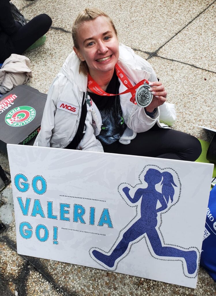 Valeria with a finisher's medal and a sign that says "Go, Valeria, go!" after a race, sitting on the ground after running